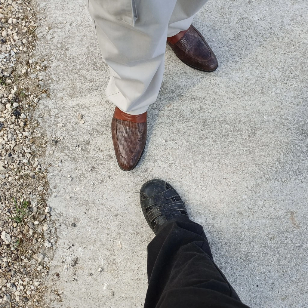 Two legs with brown shoes facing one leg with a black sandal.